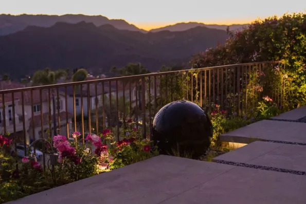 Concrete terrace with prefabricated black sphere water feature, planted railing bed, and landscape lighting overlooking hillside views at dusk in Pacific Palisades, CA