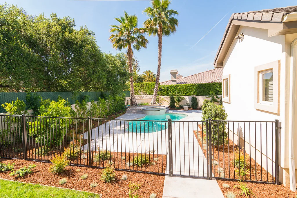 Modern backyard with concrete pool decking, fresh planting beds, mulch, drip irrigation, and wrought iron fencing.