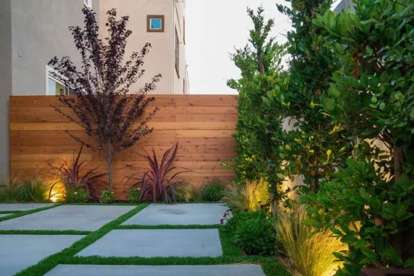 Backyard landscape with a horizontal redwood fence stained in cedar, concrete patio pads with artificial turf joints, layered planting, and warm accent lighting in Inglewood, CA.
