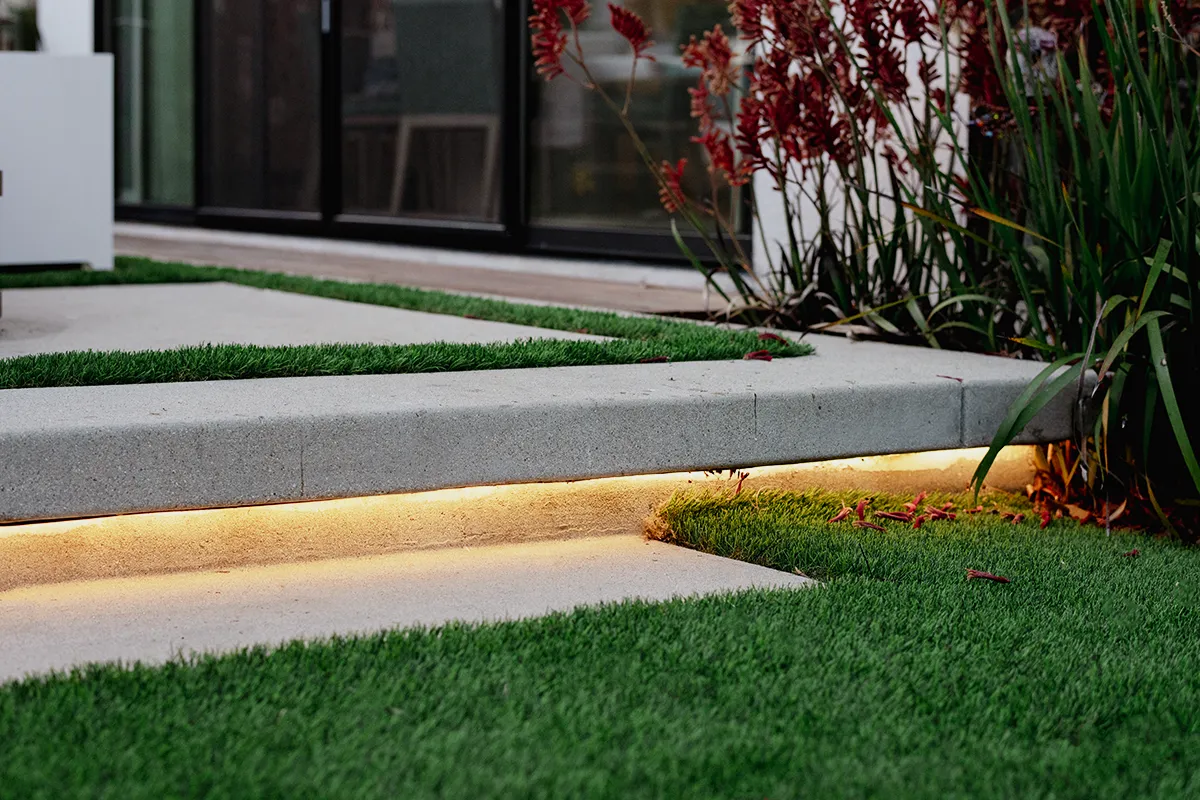 Modern concrete step with integrated LED landscape lighting, bordered by artificial turf and contemporary planting, designed and installed by Lush Gardens Inc.