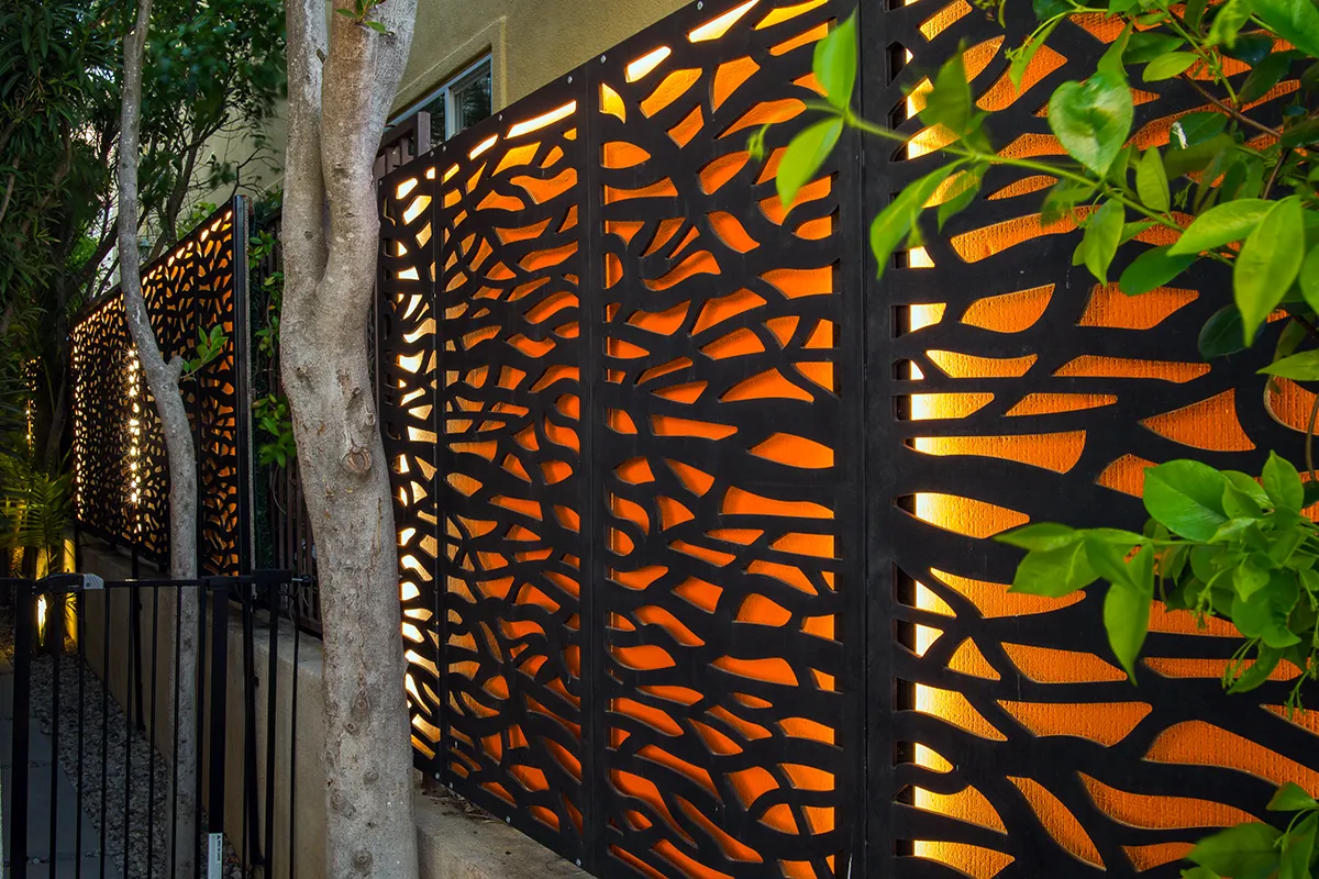 Decorative metal privacy screen with integrated warm accent landscape lighting highlighting textured panels and surrounding trees, designed and installed by Lush Gardens Inc.