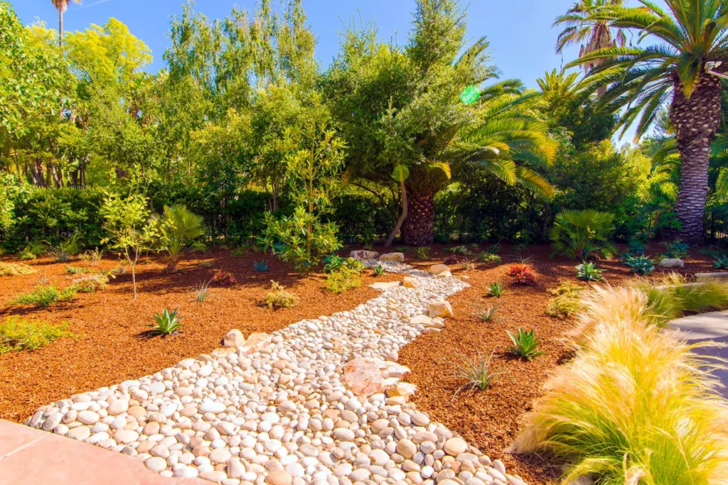 Landscape feature with a dry creek bed made of smooth river rock, mulched planting areas, drought-tolerant plants, and mature trees installed by Lush Gardens Inc.