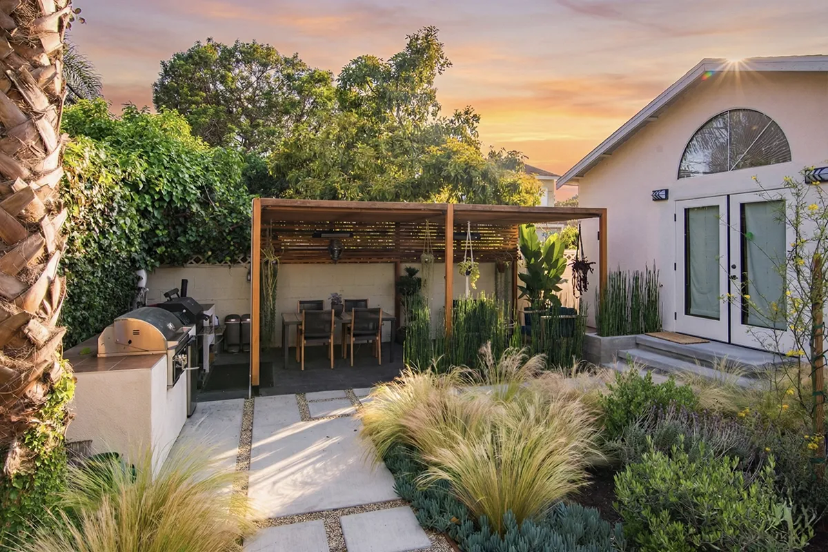 Backyard landscape construction featuring a concrete patio with stepping stone insets, wood pergola with dining area, outdoor kitchen element, ornamental grasses, layered planting beds, and integrated landscape layout by Lush Gardens Inc.