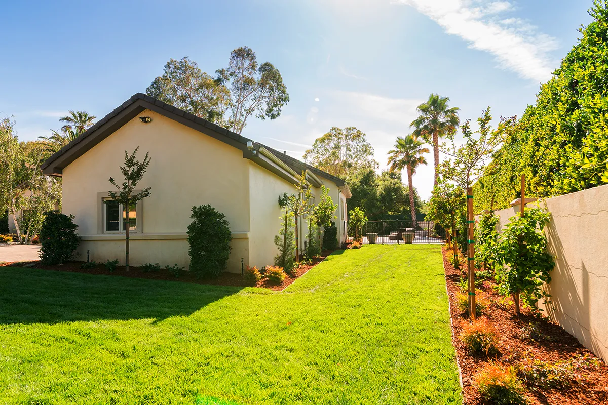 Side yard landscape construction with green lawn, mulched planting beds, young trees, shrubs, and perimeter fencing
