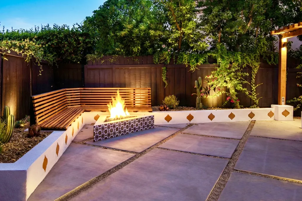 Modern backyard hardscape featuring large concrete pads with gravel joints, a custom built-in bench, decorative planter walls, and a tiled fire feature, designed and installed by Lush Gardens Inc.