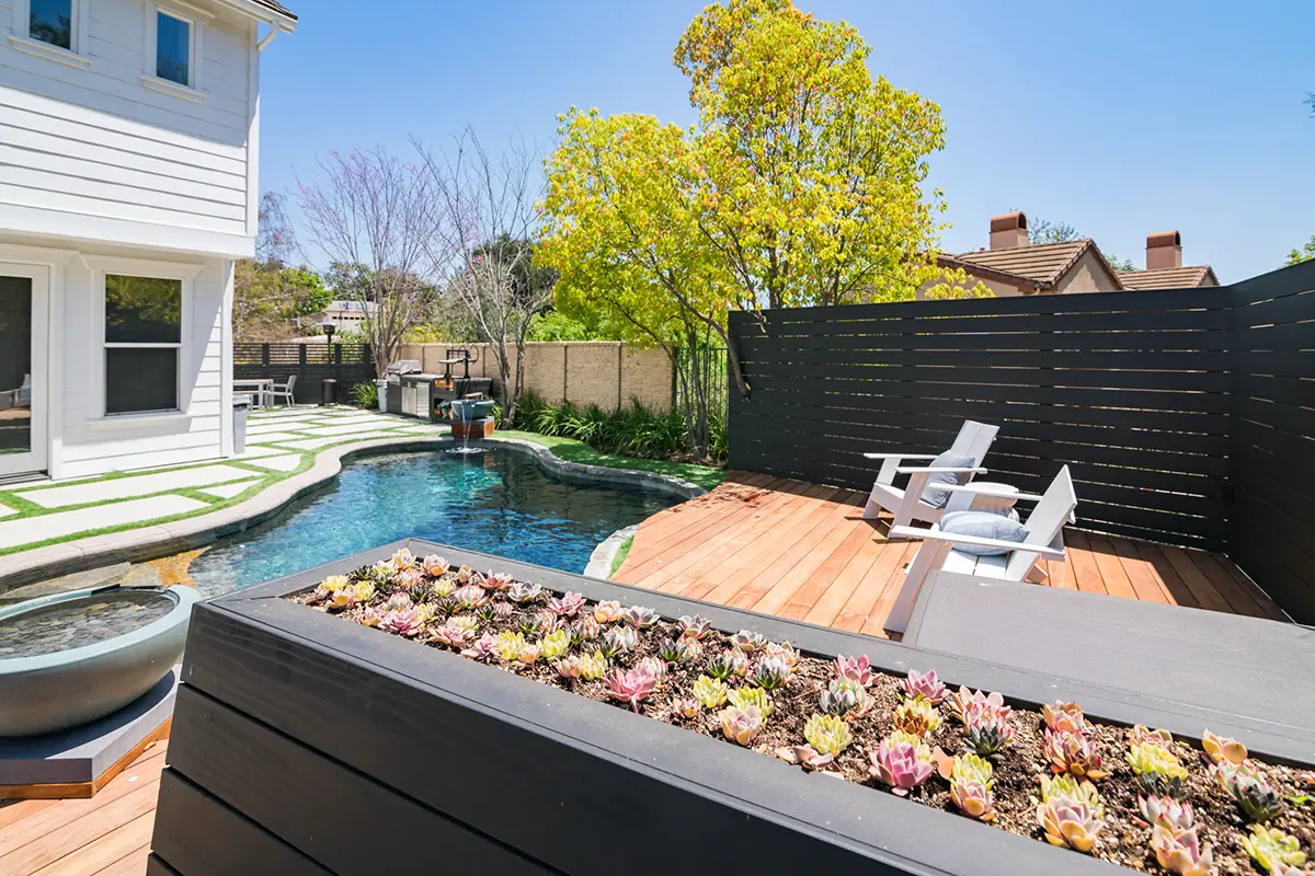 Modern backyard landscape and softscape featuring custom pool, wood deck seating area, raised planter with succulents, lawn inlay walkways, and privacy fence designed and installed by Lush Gardens Inc.