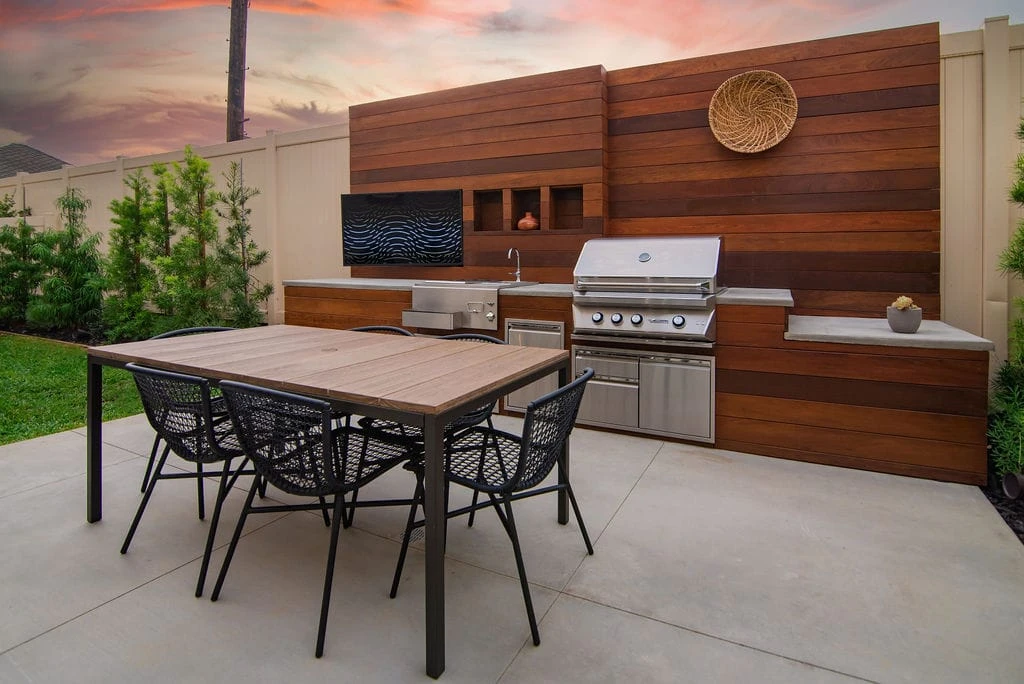 Backyard concrete patio with outdoor kitchen featuring stainless steel grill, sink, concrete countertops, decorative IPE wood wall with outdoor TV, and dining table in Inglewood, CA.