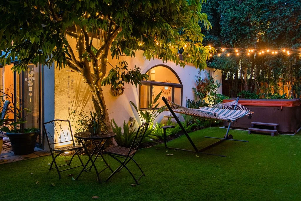 Backyard artificial turf lawn with outdoor seating, hammock, mulched planting beds, and warm landscape and string lighting in Los Angeles, CA.