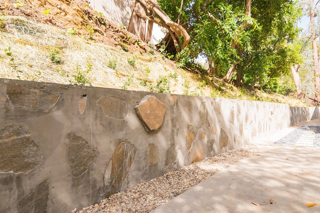 Concrete retaining wall with decorative flagstone facing, gravel edging, and planted hillside above along a concrete walkway in Hollywood Hills, Los Angeles, CA