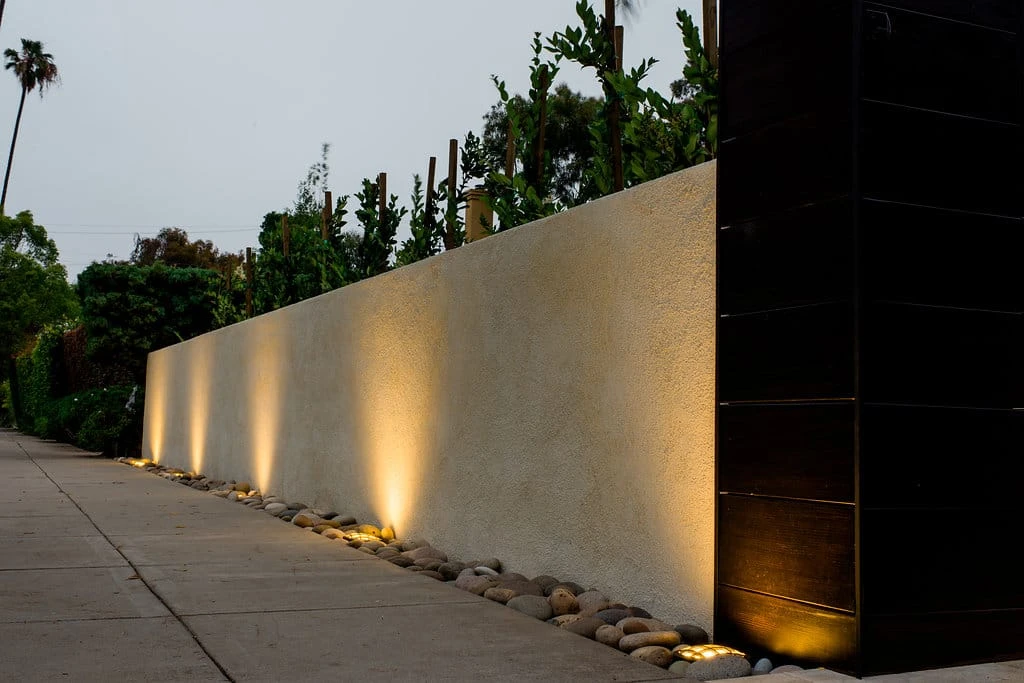Stucco-finished block wall with integrated hardscape uplighting and river rock base along concrete walkway in Los Angeles CA